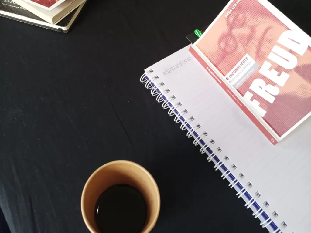 Livro de Freud e caderno de anotações sobre mesa preta, representando o estudo aprofundado na Escola de Psicanálise do Nuamce em Campinas.
