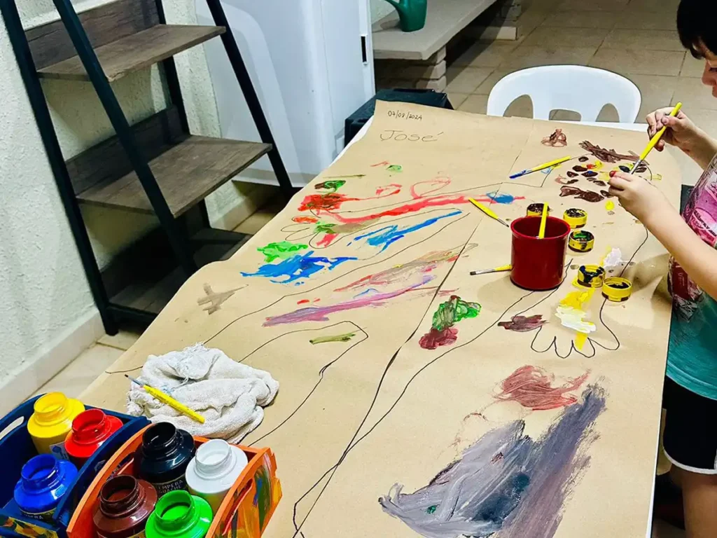Criança pintando com tinta guache colorida sobre papel kraft grande em mesa do espaço de recreação e arte terapêutica do Nuamce em Campinas.