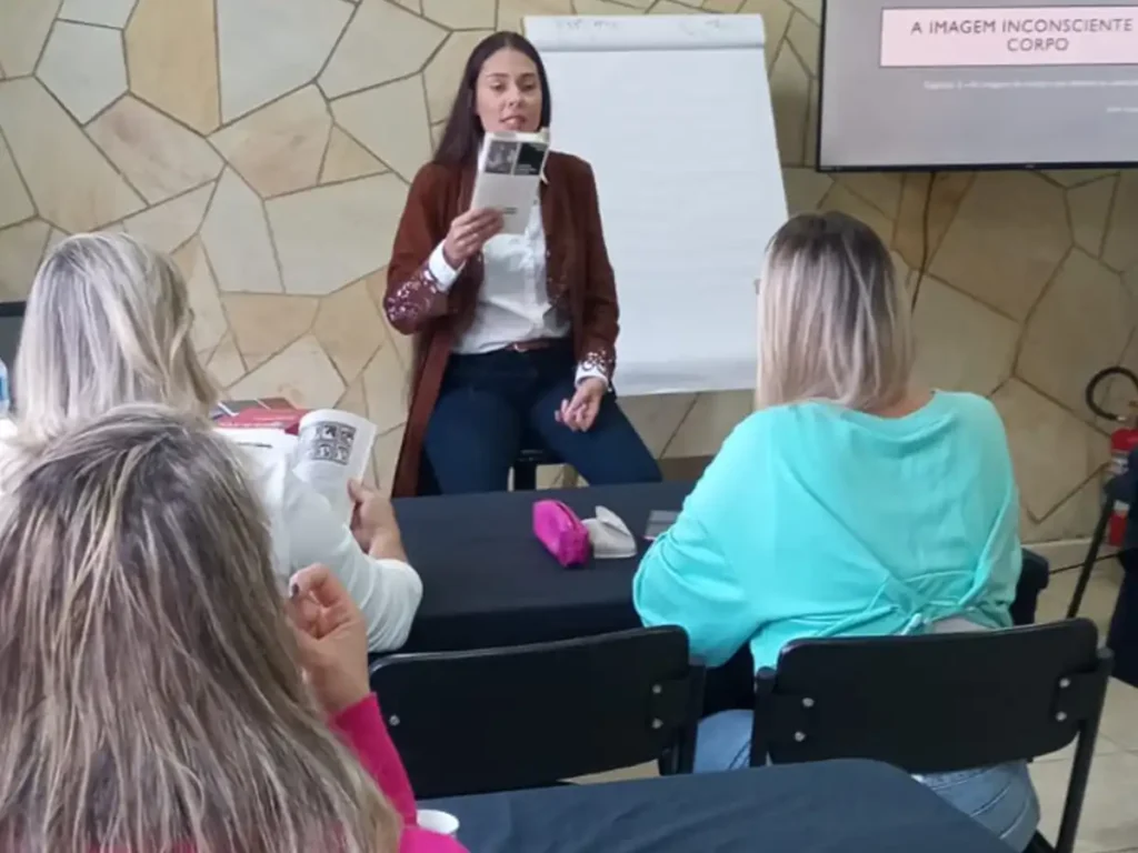 Professora em pé segurando um livro e ministrando aula para grupo de alunas durante curso de formação em psicanálise em Campinas. Ao fundo, projeção com o tema "A Imagem Inconsciente Corpo".