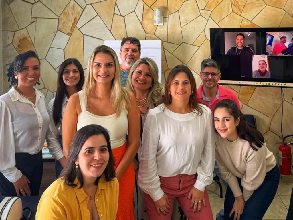 Grupo de profissionais da saúde mental reunidos após treinamento presencial em sala locada em Campinas, com participantes remotos interagindo via videoconferência na TV ao fundo.
