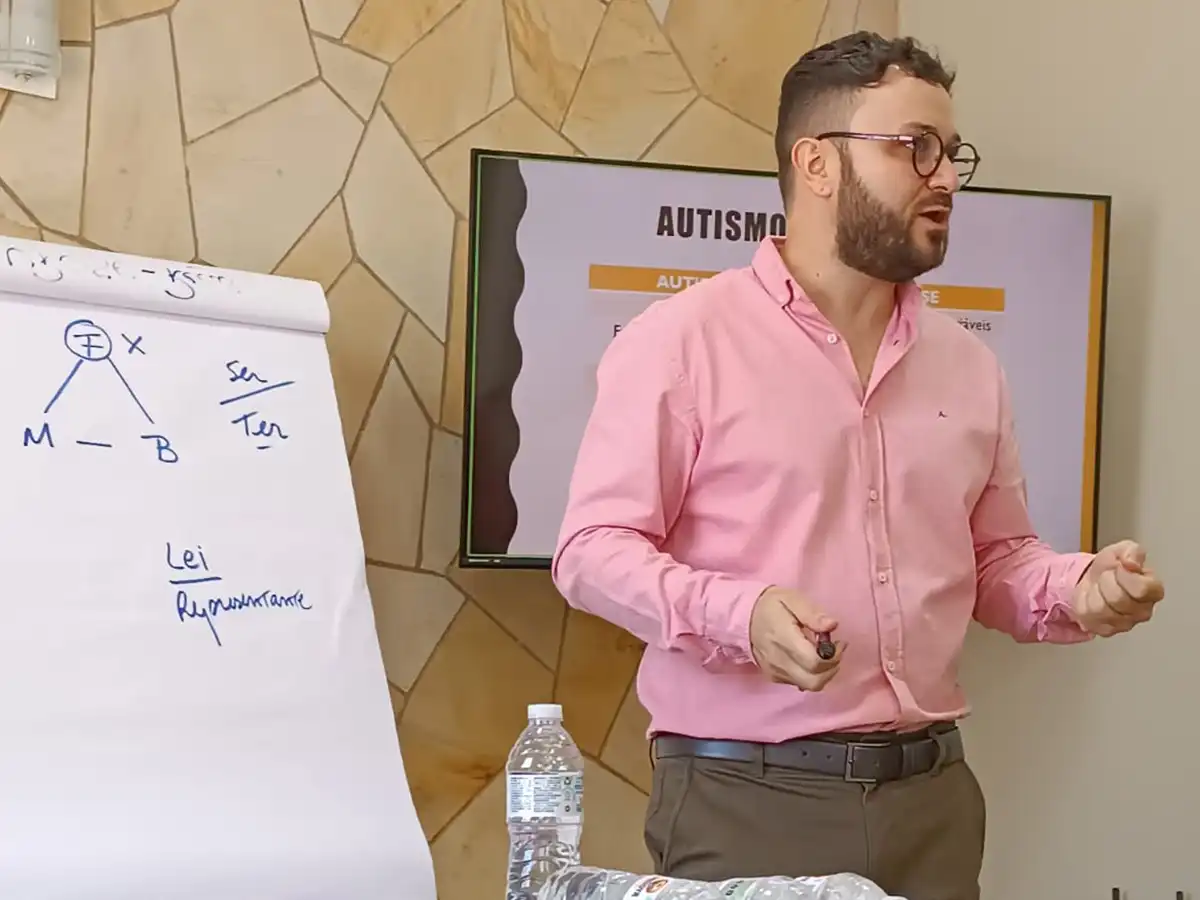 Palestrante em pé ao lado de um flip chart com esquemas psicanalíticos e uma tela com a palavra Autismo, durante palestra na Clínica Nuamce em Campinas.